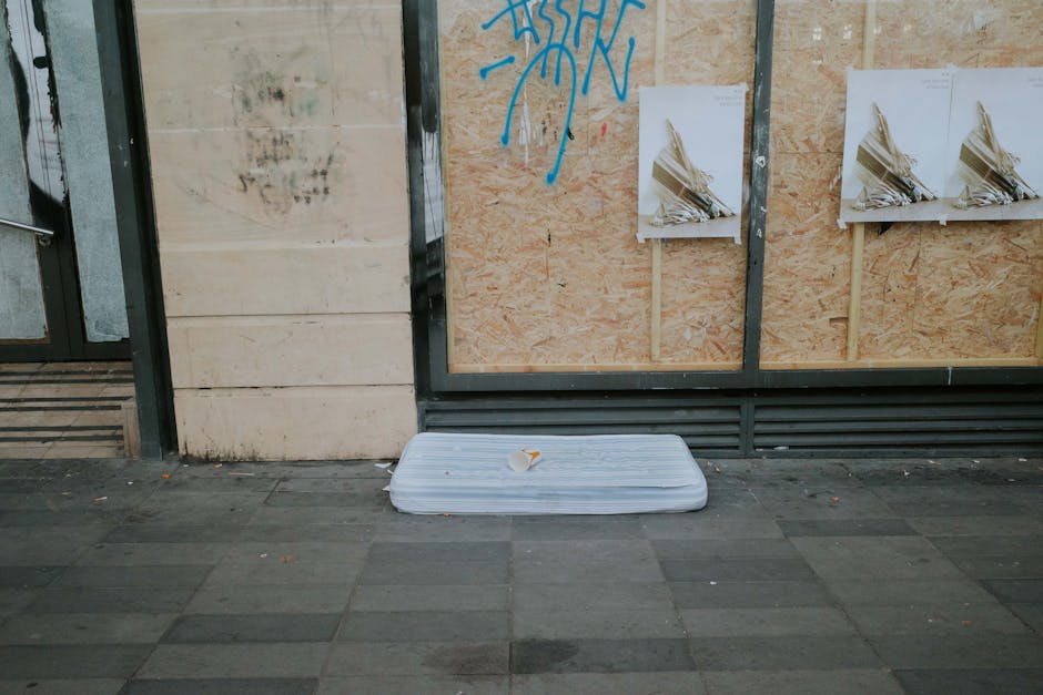 An outdoor urban scene showing a discarded, flattened white mattress leaning against a plywood board on a pavement surface. The plywood board is part of a building under construction or renovation, wi