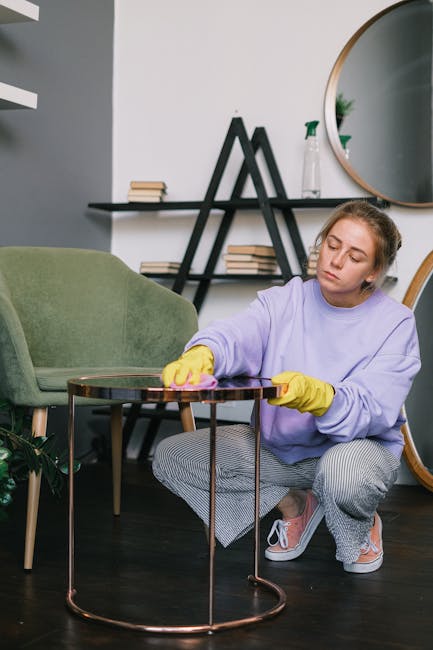 A person's hand holding a vacuum cleaner hose is seen vacuuming a decorative yellow and white patterned cushion placed on a dark fabric sofa in a living room. The sofa’s surface appears clean and well-maintained, with a neutral-colored wall in the background. The scene illustrates surface cleaning and deep cleaning processes for domestic furniture, consistent with the services offered by Enfield Carpet Cleaners, as referenced in the page about same day upholstery cleaning in Ponders End Brimsdown.